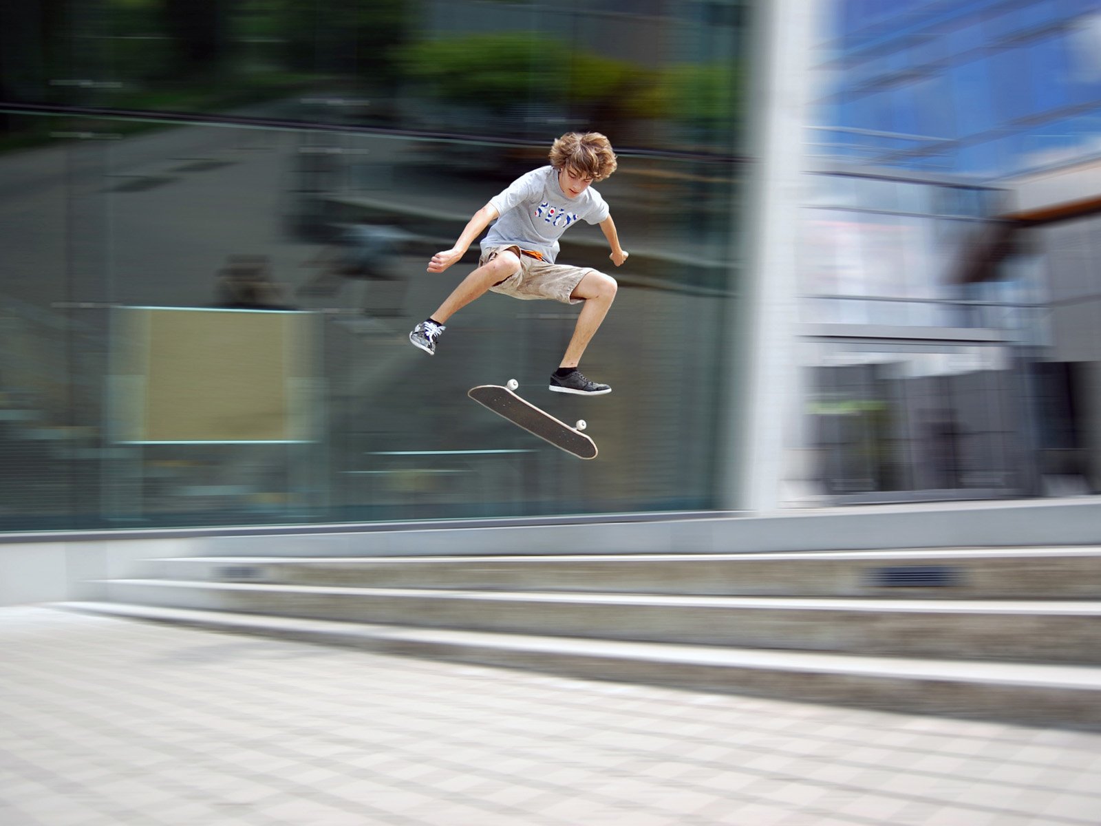 A young skateboarder performs an impressive trick mid-air in front of a modern building, capturing the dynamic energy of skateboarding and sports in this HD PC desktop wallpaper.