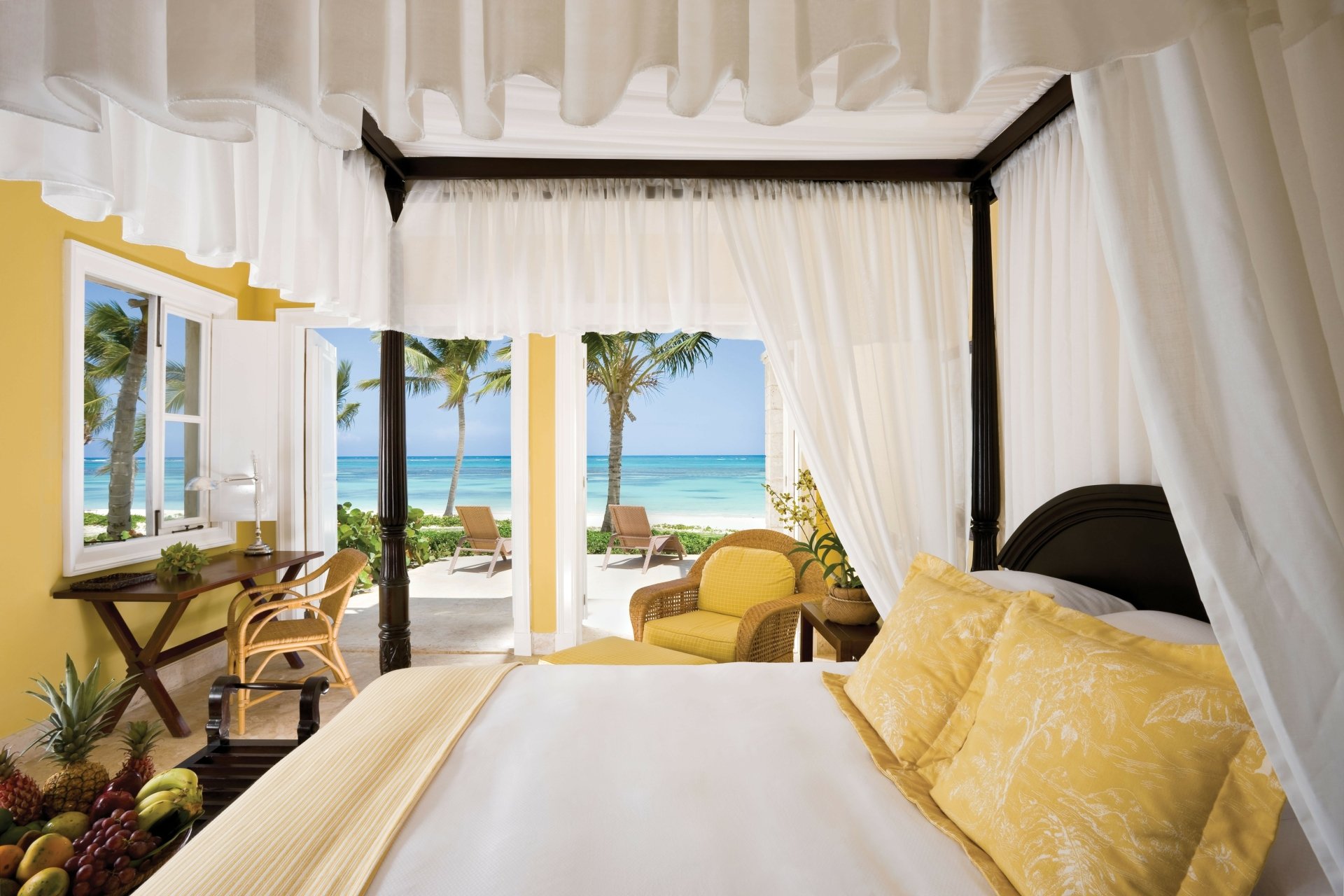 5K Ultra HD PC desktop wallpaper of a man-made tropical resort room: canopy bed with yellow linens opening to a palm-lined beach and turquoise ocean.