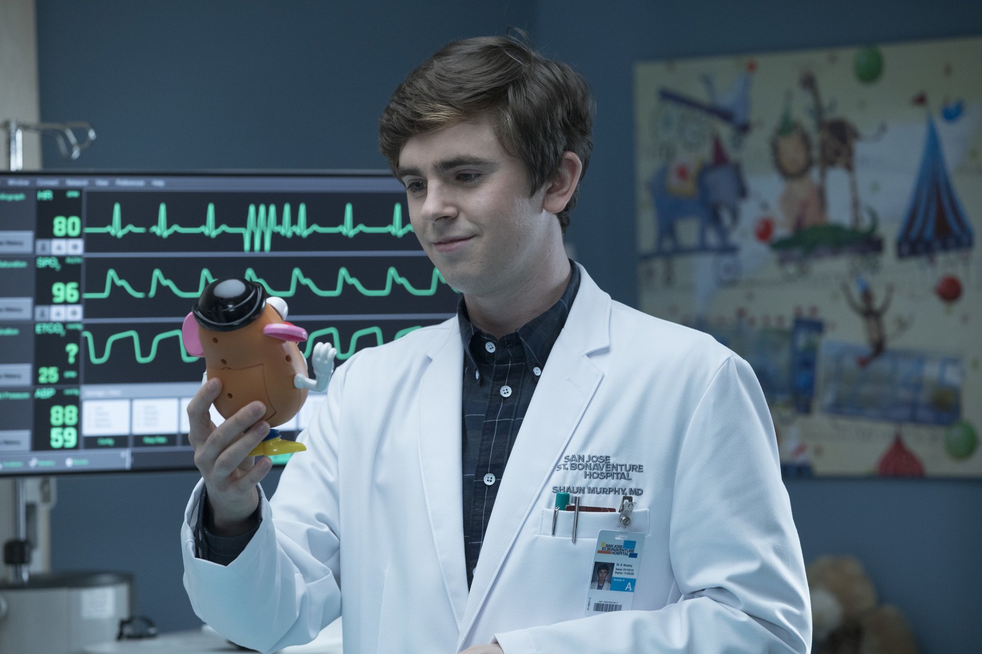 HD wallpaper featuring Freddie Highmore from The Good Doctor, in a lab coat, examining a toy in a clinical setting.