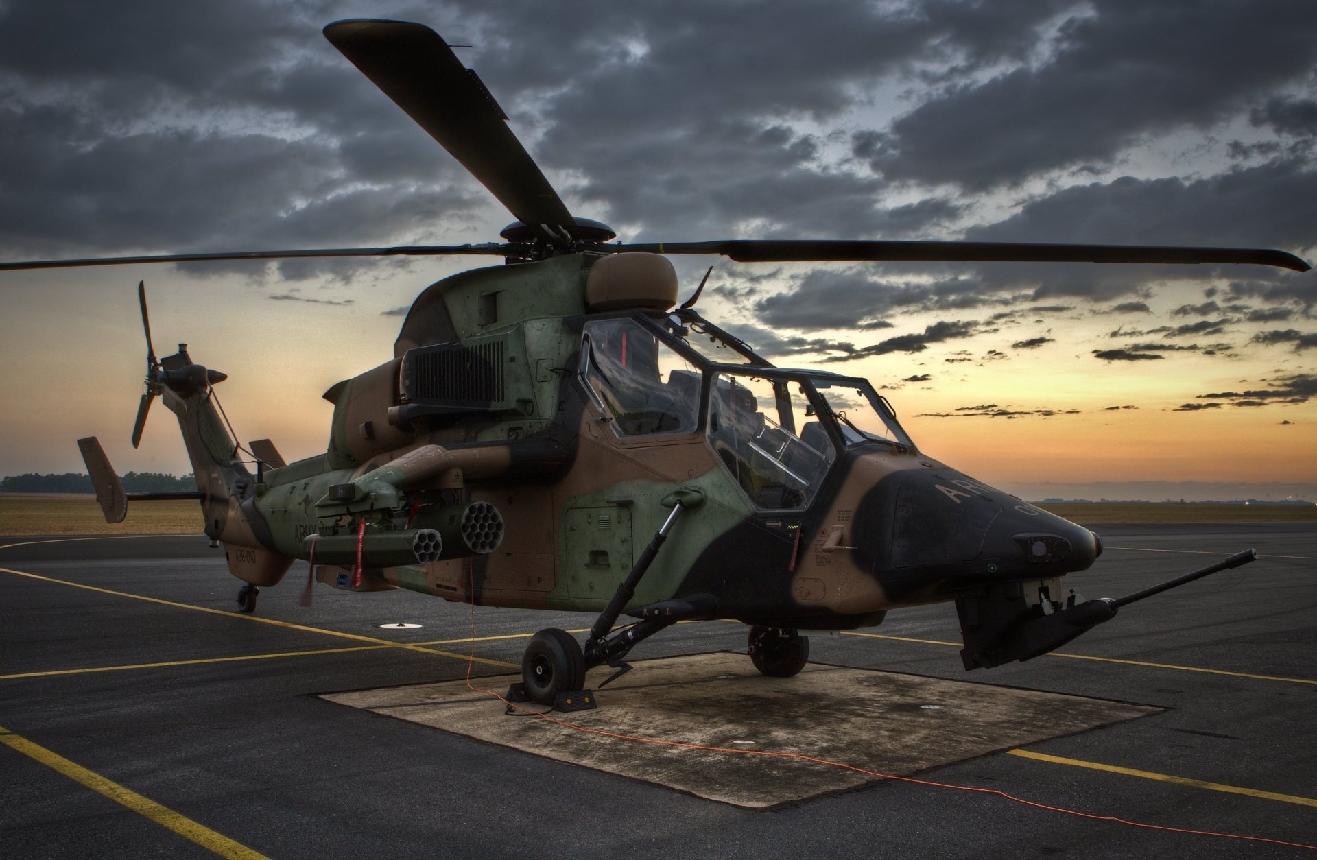 HD desktop wallpaper featuring a Eurocopter Tiger attack helicopter on a military airfield at dusk under a dramatic sky.