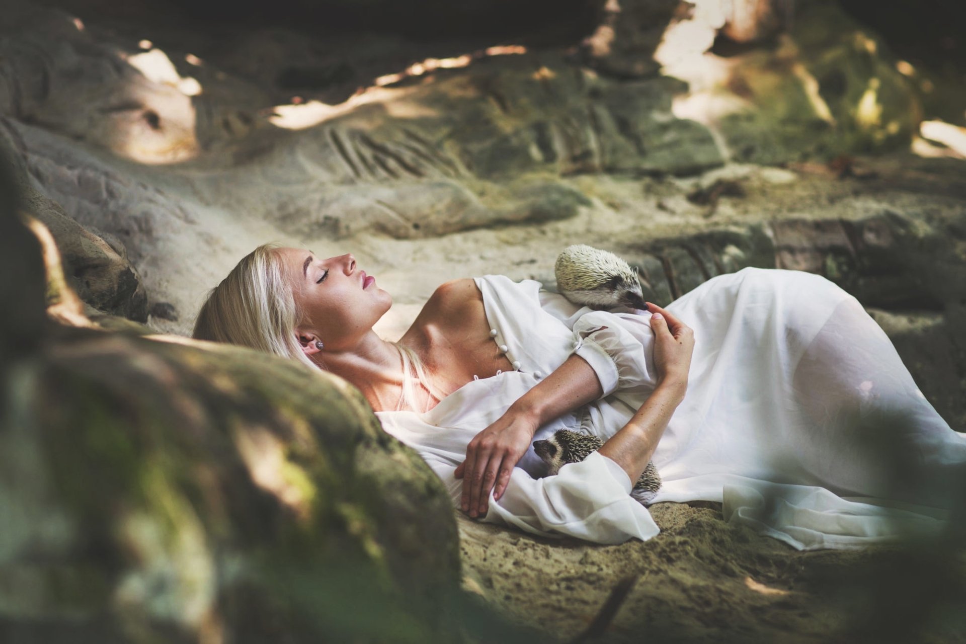 A blonde woman in a white dress lies peacefully on rocky ground, gently holding a small hedgehog, evoking a serene and natural mood in this HD desktop wallpaper.