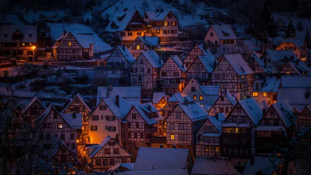 HD wallpaper of a snow-covered village at night, showcasing warmly lit, quaint houses under a serene blue twilight.