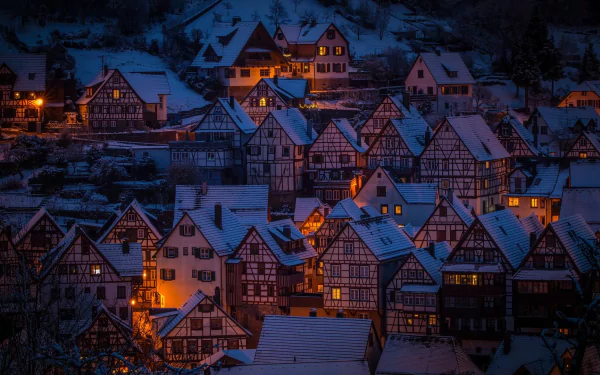 HD wallpaper of a snow-covered village at night, showcasing warmly lit, quaint houses under a serene blue twilight.