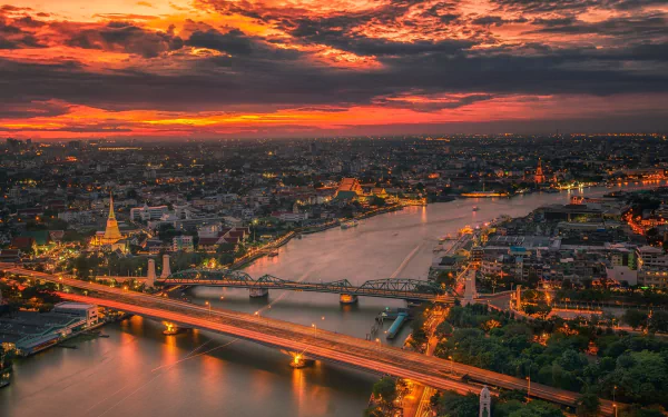 A vibrant sunset over Bangkok, Thailand, illuminating the cityscape and river with warm cloud-filled skies and glowing urban lights at night.