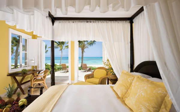 5K Ultra HD PC desktop wallpaper of a man-made tropical resort room: canopy bed with yellow linens opening to a palm-lined beach and turquoise ocean.