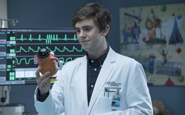 HD wallpaper featuring Freddie Highmore from The Good Doctor, in a lab coat, examining a toy in a clinical setting.