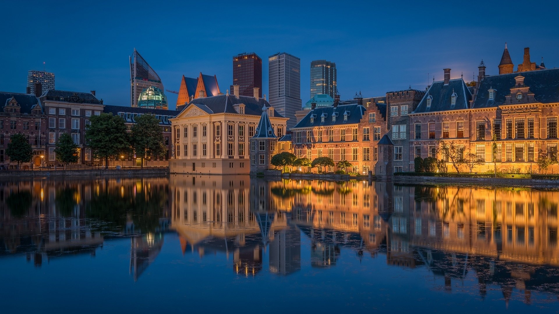 Night view of The Hague cityscape with illuminated historic and modern buildings reflecting on calm water, captured in HD for a striking desktop wallpaper.