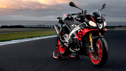 A 4K Ultra HD desktop wallpaper showcasing the Aprilia Tuono V4 1100 Factory motorcycle parked on a racetrack at sunset.