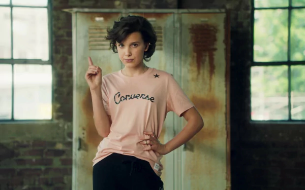 Black-haired woman in a pink tee stands by an industrial window, one hand on hip and finger raised in a model-like pose; 2K Quad HD PC desktop wallpaper/background.