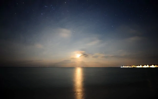 Nighttime ocean view with moonset reflecting over calm water, clouds drifting above, and hotel lights glowing along the distant shore under a serene sunset sky.
