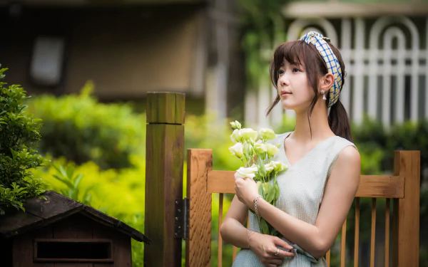 An Asian brunette woman in a dress holds white flowers, seated outdoors with blurred greenery and wooden fence in the background, captured in 8K Ultra HD.