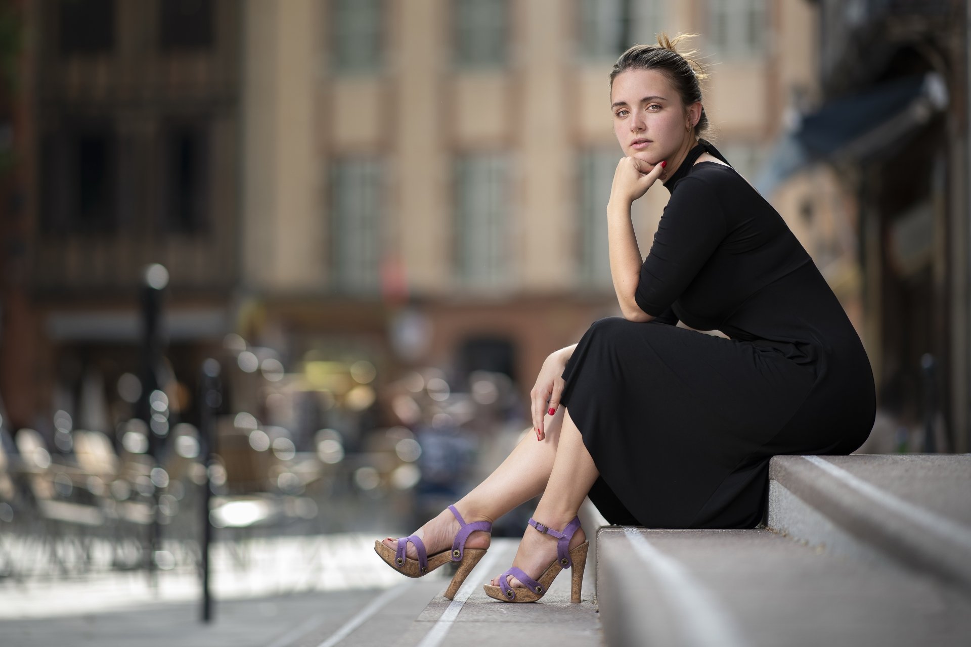 A model in a black dress and high heels sits on steps with a blurred city background, showcasing depth of field in this 4K Ultra HD desktop wallpaper.