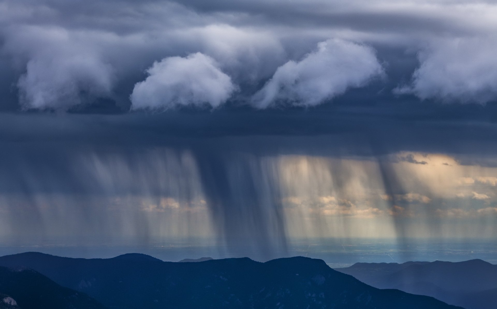 HD desktop wallpaper showing a dramatic rainstorm over mountain peaks, with dark clouds and streaks of rain descending from the sky, capturing the raw beauty and power of nature.