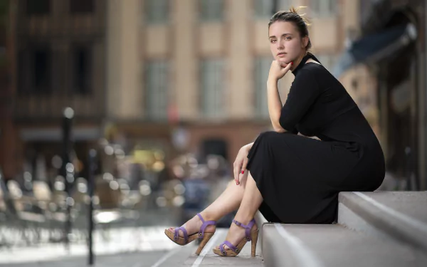 A model in a black dress and high heels sits on steps with a blurred city background, showcasing depth of field in this 4K Ultra HD desktop wallpaper.
