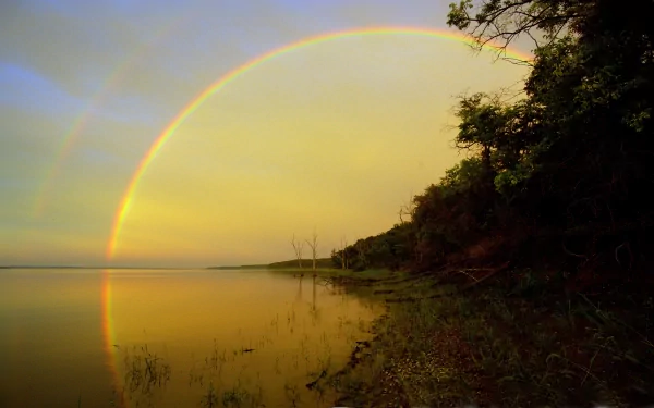 A tranquil nature scene featuring a vibrant rainbow arching over a serene lake, with lush greenery along the shoreline, showcasing the beauty of the outdoors. HD desktop wallpaper background.