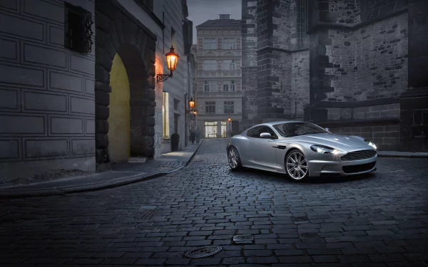 Silver Aston Martin DBS supercar parked on a cobblestone street in a moody cityscape — 4K Ultra HD PC desktop wallpaper background.