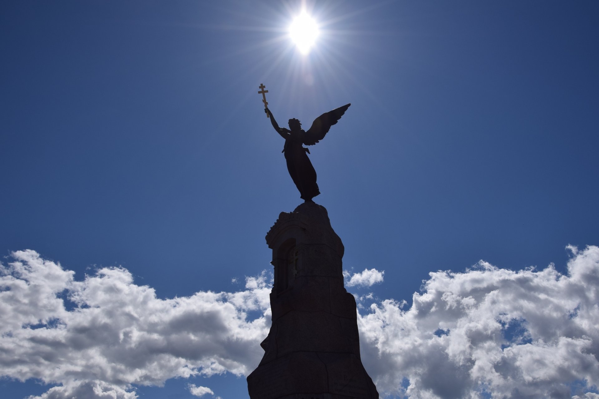 Silhouette of an angel statue on a monument with a sunbeam shining above, set against a blue sky with scattered clouds, captured in 4K Ultra HD.