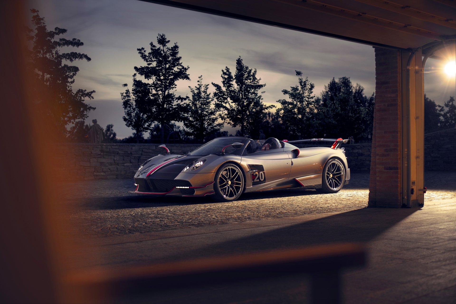 8K Ultra HD PC desktop wallpaper of a silver Pagani Huayra supercar parked at dusk, a low-slung vehicle framed by a garage opening with silhouetted trees in the background.