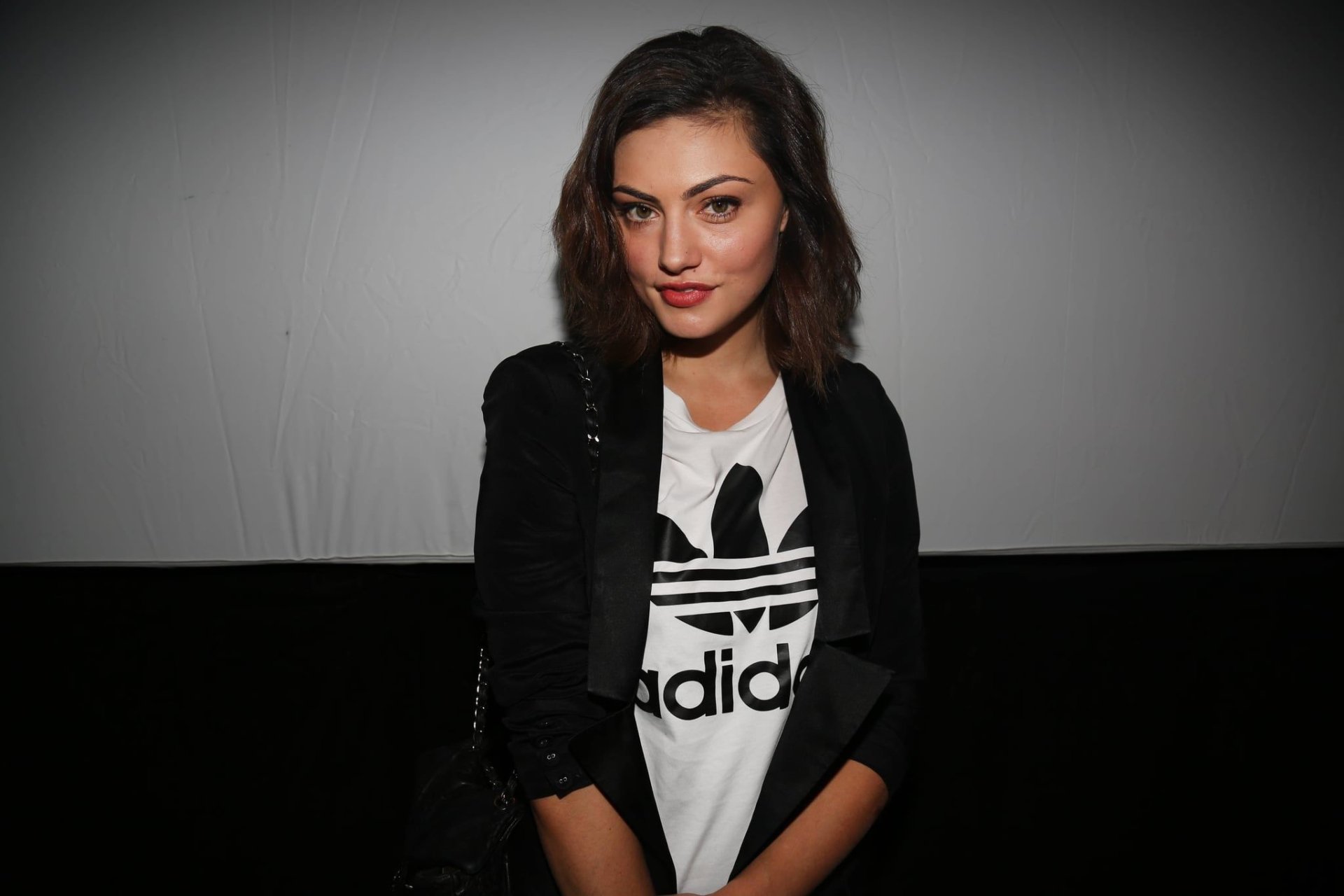 Brunette woman in a black jacket over a white Adidas tee, seated against a gray-to-black backdrop, framed as an HD PC desktop wallpaper.