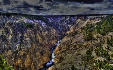 An HDR image of Yellowstone National Park featuring a dramatic canyon with a river running through it, surrounded by rugged cliffs and sparse trees under a cloudy sky, serving as a HD desktop wallpaper.
