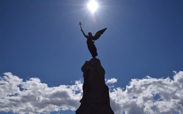 Silhouette of an angel statue on a monument with a sunbeam shining above, set against a blue sky with scattered clouds, captured in 4K Ultra HD.