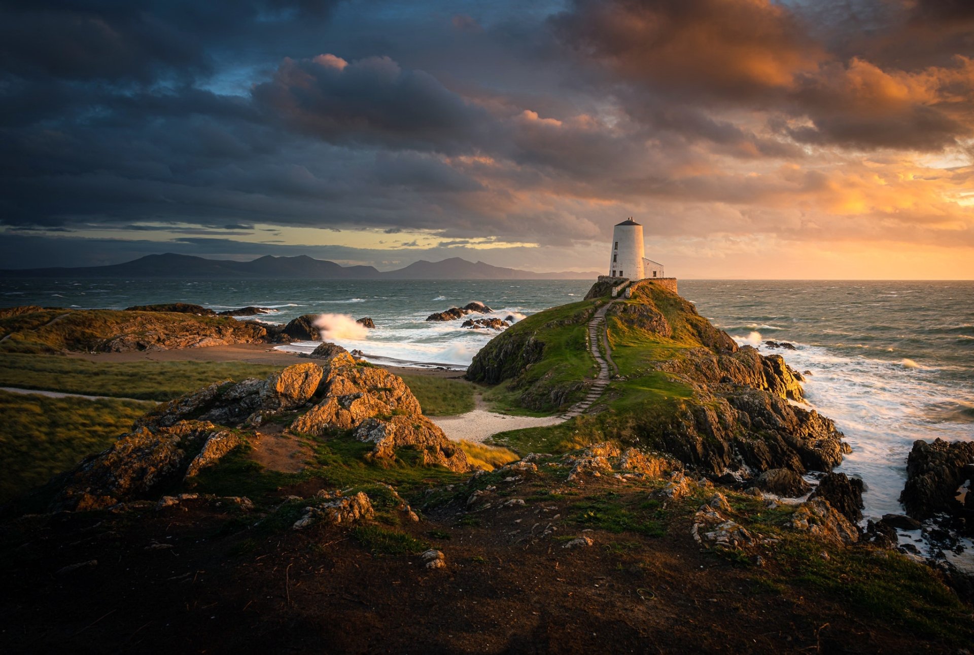 A man-made lighthouse stands on a rocky path along the Welsh coast, overlooking the sea and horizon under a dramatic, colorful sky.