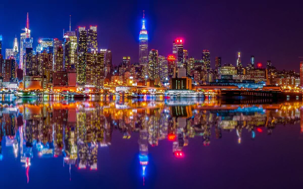 Nighttime view of Manhattan skyscrapers along the Hudson River in New York, with vibrant city lights reflecting on the water, captured in a high-definition desktop wallpaper.