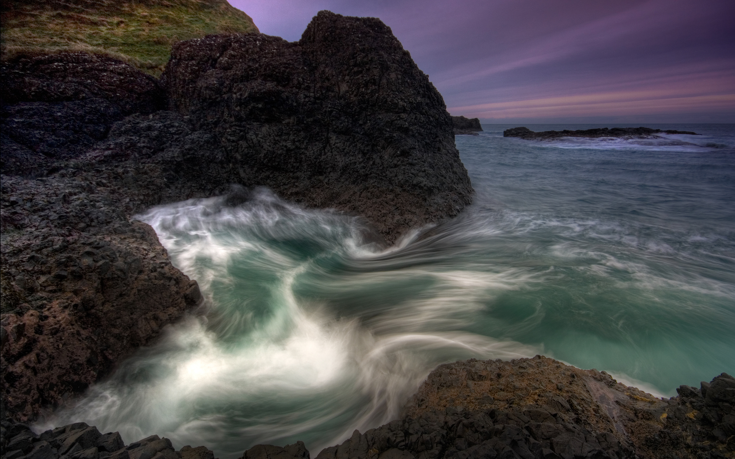 Coastal sunset in Ireland with a rushing wave crashing against the shore at dusk.