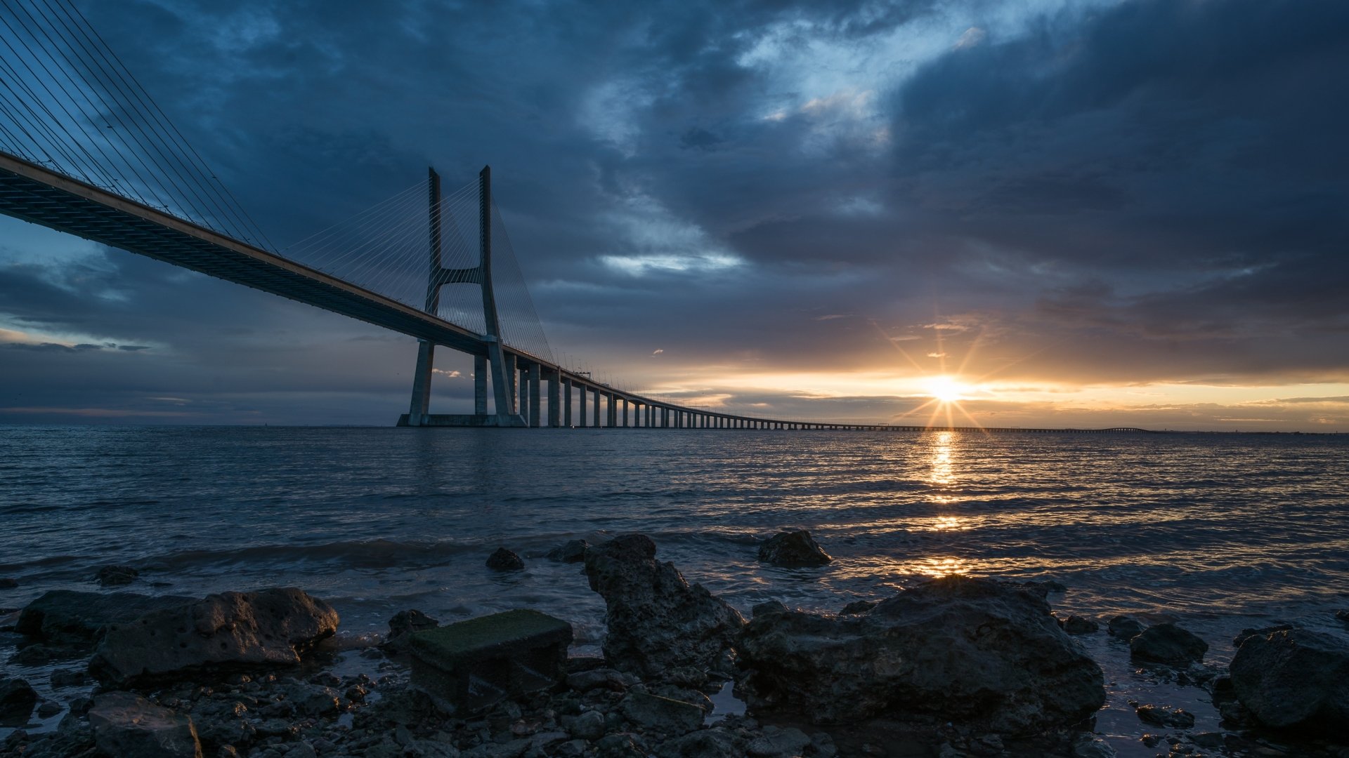 Sunrise over the Vasco da Gama Bridge in Portugal, with dramatic clouds and calm waters on the horizon, captured as an HD desktop wallpaper background.