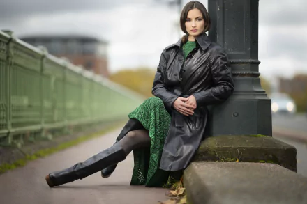 Brunette model in a leather jacket seated by a lamppost against a blurred city backdrop; pronounced depth of field, 5K Ultra HD PC desktop wallpaper and background.