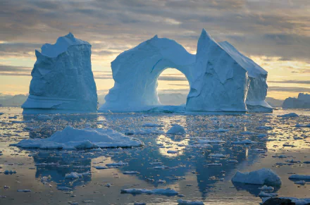 HD desktop wallpaper showing a natural iceberg arch with its reflection on calm water, surrounded by floating ice under a cloudy sky.