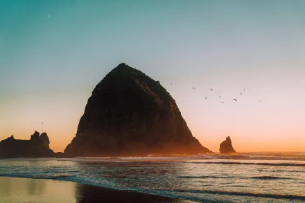 4K Ultra HD PC desktop wallpaper: Haystack Rock at Cannon Beach, Oregon — a towering sea stack at sunset, waves on the sandy beach and birds in a pastel sky.