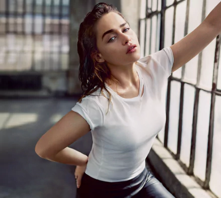 HD wallpaper of English actress and celebrity Emilia Clarke. She is shown with blue eyes, brunette hair, and wearing a white shirt, posing by large windows.