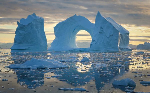 HD desktop wallpaper showing a natural iceberg arch with its reflection on calm water, surrounded by floating ice under a cloudy sky.