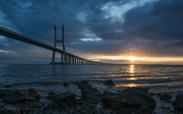 Sunrise over the Vasco da Gama Bridge in Portugal, with dramatic clouds and calm waters on the horizon, captured as an HD desktop wallpaper background.