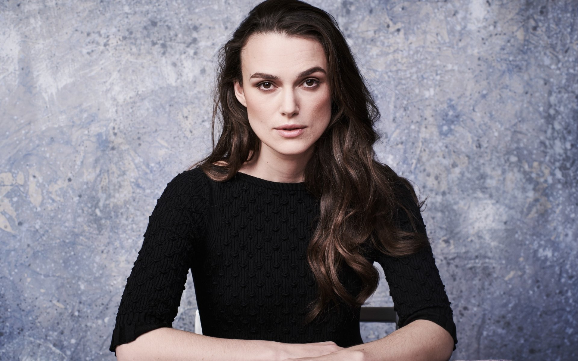 2K Quad HD PC desktop wallpaper: portrait of a brunette woman with long brown hair and brown eyes, wearing a black top, seated against a textured gray backdrop.