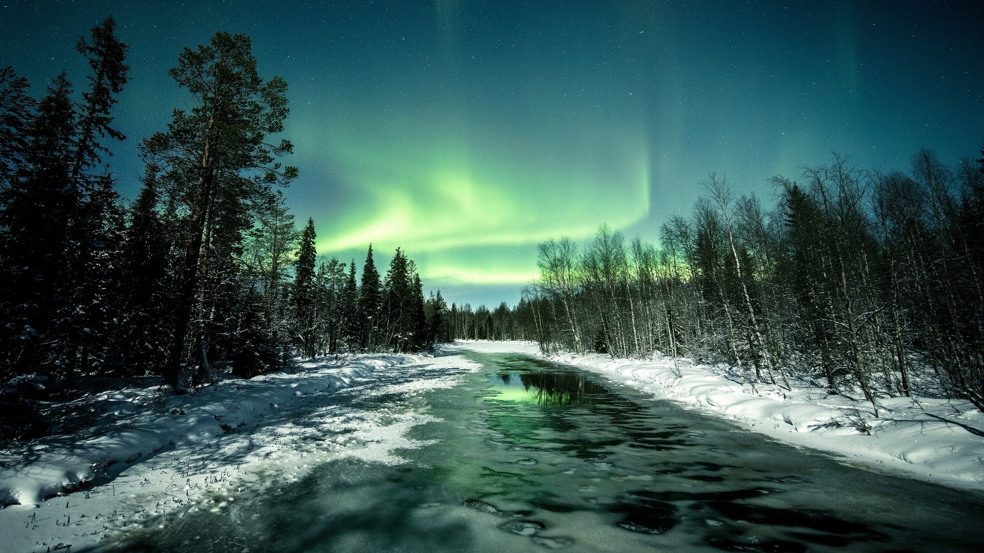 A serene winter night by a snowy river under a starry sky, illuminated by vibrant green aurora borealis in this 4K Ultra HD nature wallpaper.