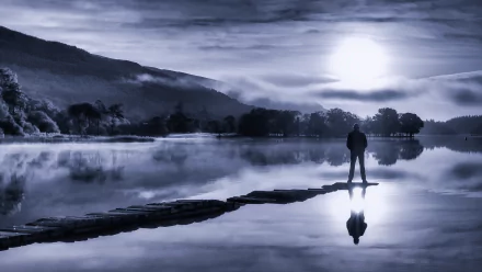 Monochrome 4K Ultra HD wallpaper of a man standing alone on a rock path at sunset, reflected in a calm lake with mist and silhouetted trees in the background.