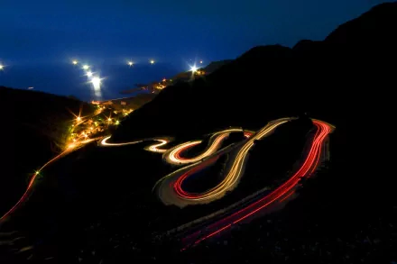 A mesmerizing time-lapse photograph capturing the light trails of cars winding along a coastal road, set against a serene night backdrop, creating a stunning HD desktop wallpaper.