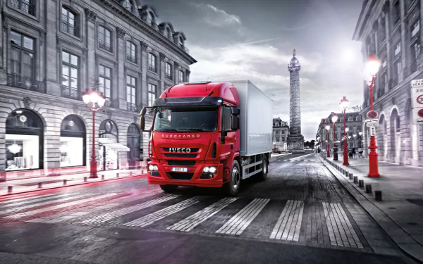 HD PC desktop wallpaper featuring a red Iveco 180e32 truck driving through a city street with classic buildings and a tall monument in the background.