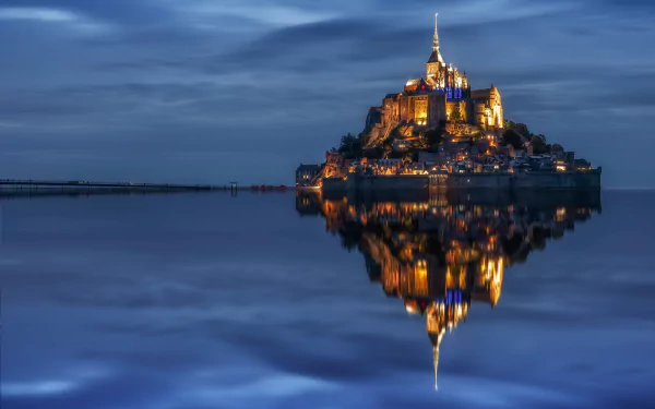 Mont Saint-Michel monastery illuminated at night, reflecting perfectly on the calm water in France, captured as an HD desktop wallpaper and background.