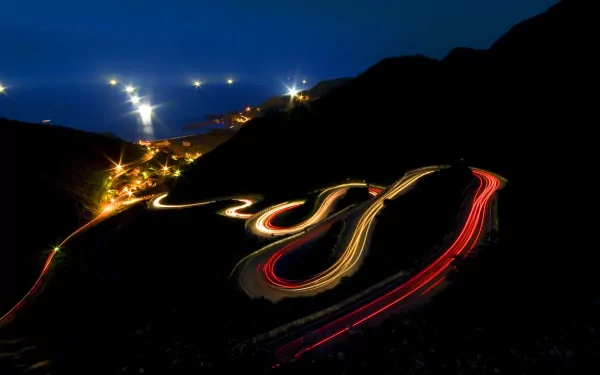 A mesmerizing time-lapse photograph capturing the light trails of cars winding along a coastal road, set against a serene night backdrop, creating a stunning HD desktop wallpaper.