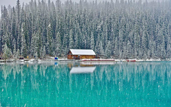 HD desktop wallpaper of a wooden cabin by a turquoise lake, with a snowy forest reflected in the water.