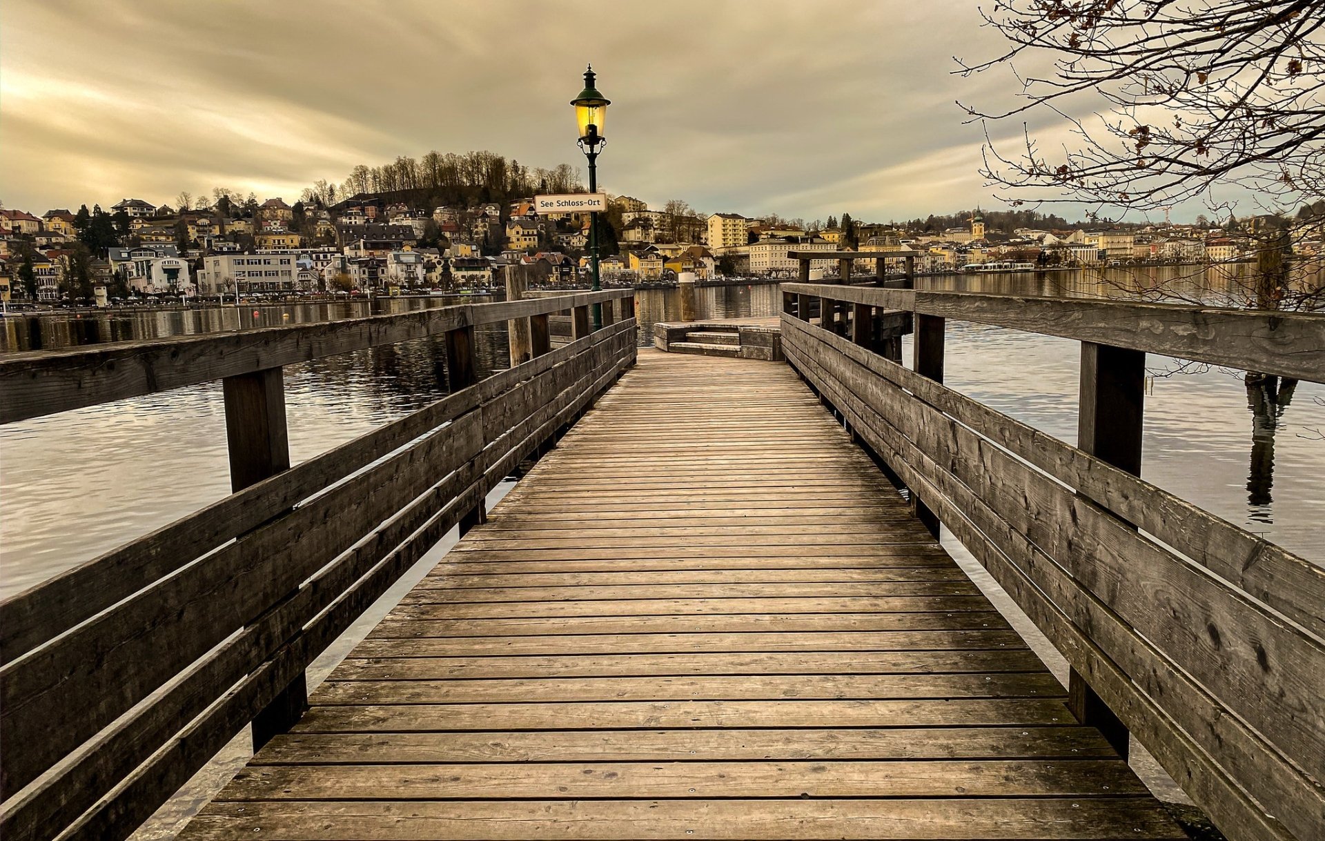 HD PC desktop wallpaper: wooden pier in Austria leading to a lamp post, a man-made walkway over a calm river with town and hills beyond.