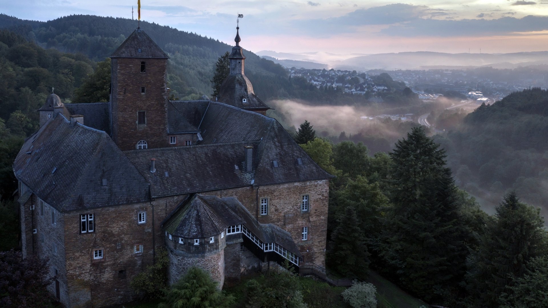 4K Ultra HD desktop wallpaper featuring a historic man-made castle perched on a hillside overlooking a misty forested valley at dawn.