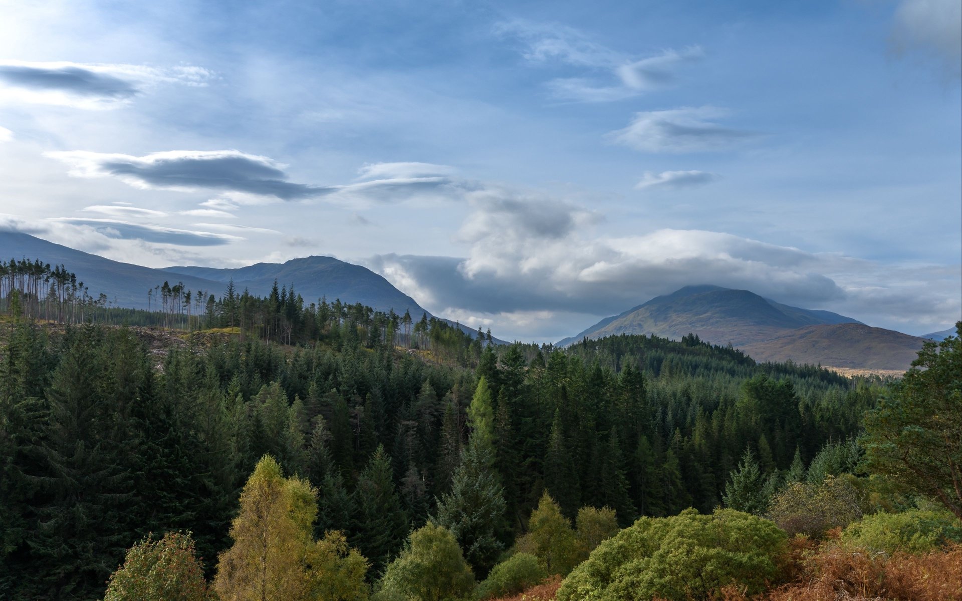 4K Ultra HD PC desktop wallpaper: panoramic mountain nature landscape with layered forested hills, mist in valleys, and a blue sky with wispy clouds.