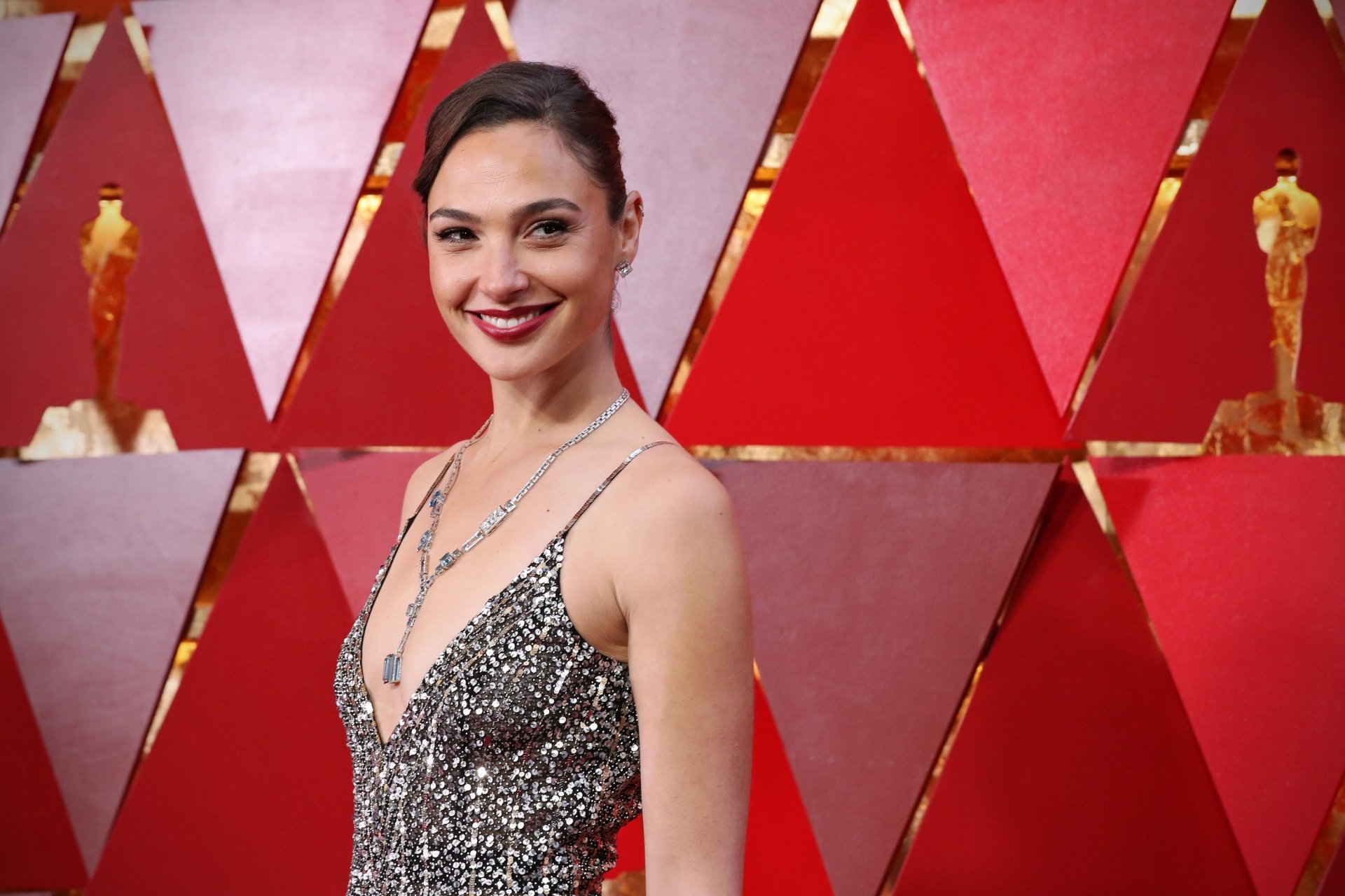 HD desktop wallpaper featuring Israeli actress and celebrity Gal Gadot with brunette hair, red lipstick, and a radiant smile against a red geometric backdrop.