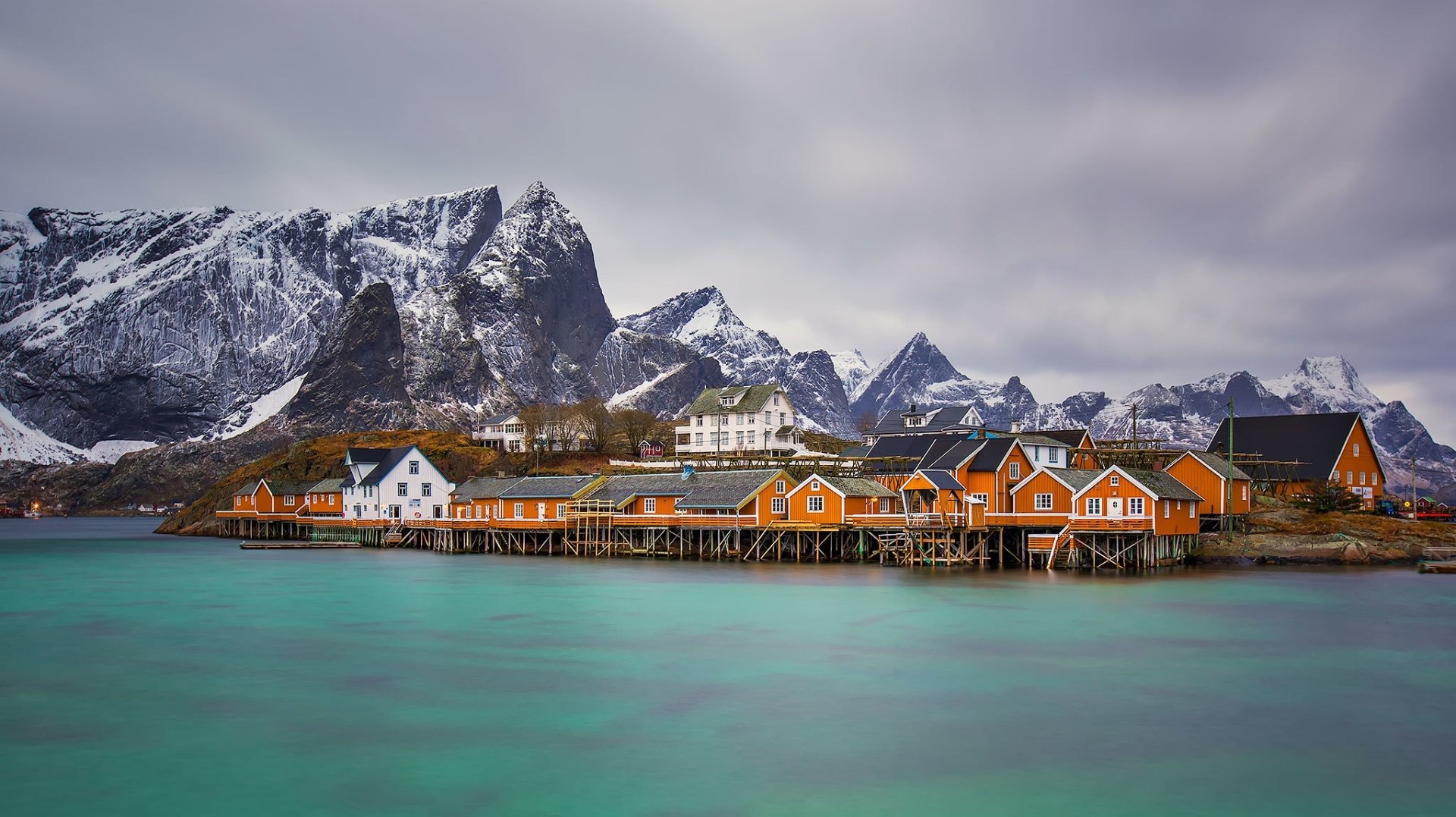 Scenic view of Reine village with colorful houses on stilts along turquoise waters, set against snowy mountains in the Lofoten Islands, Norway.