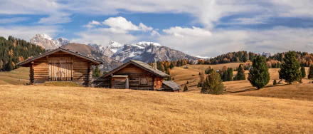 4K Ultra HD wallpaper showing man-made wooden cabins set against the fall-colored Dolomites under a partly cloudy sky.