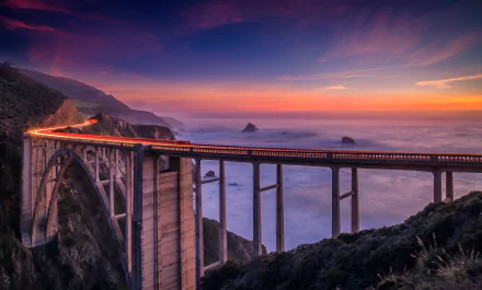 HD wallpaper of Bixby Creek Bridge on California's coast at sunset, with mountains, ocean fog, and a vibrant sky creating a picturesque landscape.